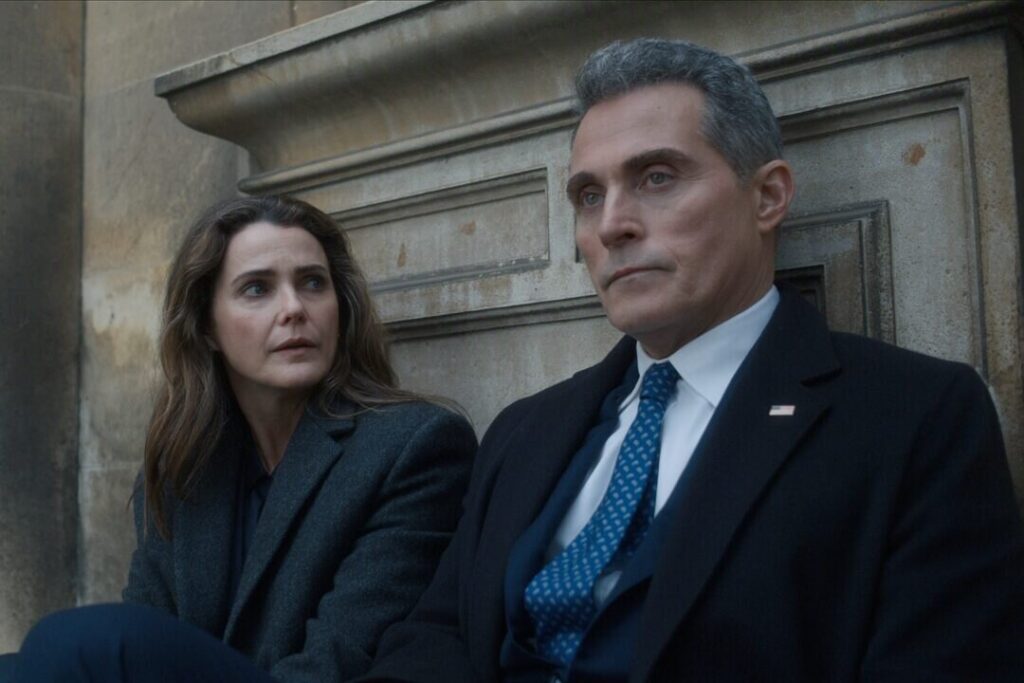 "Two people in formal coats sit on outdoor stone steps, appearing serious and deep in conversation, with an old stone wall in the background. The man wears a suit and tie with an American flag pin, suggesting a political or official context."